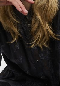 Blont hår över mörk mönstrad knappad skjorta, hand nära hakan med naturliga naglar, inget synligt ansikte eller bakgrund.