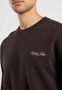 Felpa marrone con scollo rotondo e polsini a coste, con "Eighty Nine" ricamato in filo chiaro sul lato sinistro del petto.