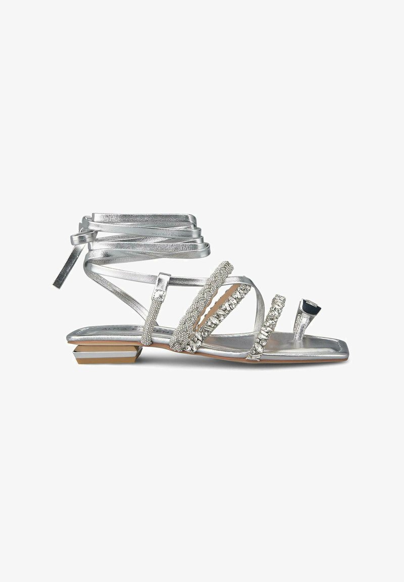 Sandale argentée à lanières avec un talon plat, ornée de perles et de strass, présentant une texture métallisée et des lanières qui se nouent autour de la cheville.