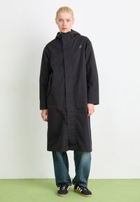 Svart vanntett jakke med hette og frontlomme, med sidelommer og rett design, kombinert med blå, litt falmede jeans og svarte joggesko.