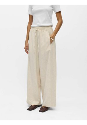 Kobieta nosząca beżowe szerokie lniane spodnie z wiązaniem w talii, biały top z krótkim rękawem oraz ciemnobrązowe buty z kwadratowym noskiem.