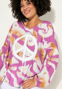 Maglietta oversize grafica in un motivo tie-dye viola, bianco e arancione. Presenta un grande segno di pace bianco centrale con gocce e dettagli in strass.