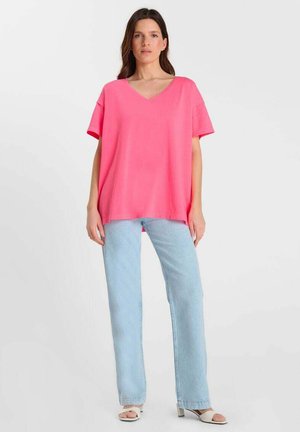 Vrouw staand, gekleed in een felroze losse T-shirt met V-hals, lichtblauwe jeans met rechte pijpen en witte open-teenhakken, tegen een effen achtergrond.