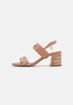 Beige sandal med åben tå og blokhæl, ankelrem med guldspænde og charm, frontrem med subtilt mønstertryk og tekstureret hældesign.