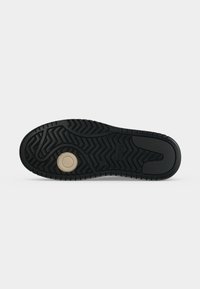 Suola nera della scarpa con motivo scolpito e dettaglio circolare beige vicino alla zona della pianta del piede su sfondo bianco.