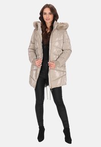 Bézs színű, fényes anyagú pufi kabát, szőrmebéléssel ellátott kapucnival, cipzáras záródással és bordázott mandzsettával. Fekete nadrággal és csizmával párosítva.