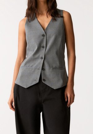 Woman wearing a sleeveless gray plaid buttoned vest paired with loose-fitting black pants against a neutral background.