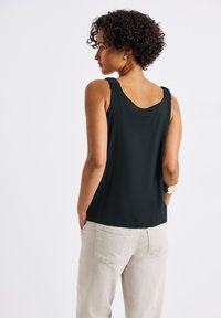 Musta toppi leveillä olkaimilla ja kaarevalla takasuunnittelulla, yhdistettynä vaaleanbeigeihin housuihin, kädet taskuissa, teksturoitu kangas.