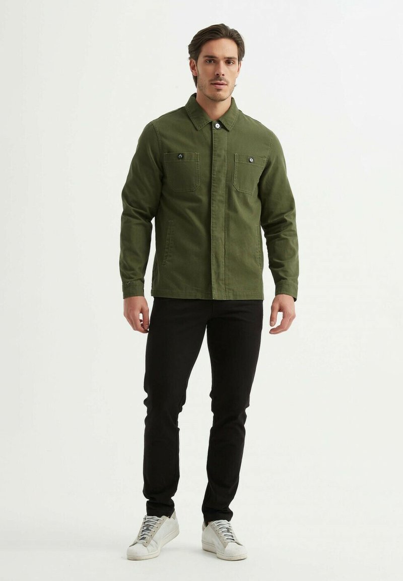 Veste en coton vert avec deux poches poitrine, fermeture à boutons et col, associée à un pantalon noir et des baskets blanches.