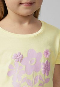 Żółta koszulka z różowymi grafikami kwiatów, z teksturowymi i błyszczącymi akcentami kwiatowymi. Krótkie rękawy i okrągły dekolt.