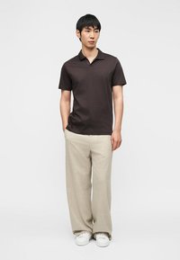 Άνδρας που στέκεται με τα χέρια στις τσέπες, φορώντας σκούρο καφέ κοντομάνικο πόλο, μπεζ παντελόνι με φαρδιά πόδια και λευκά αθλητικά σε λευκό φόντο.