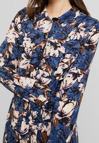 Blommig blus med blå, rosa och bruna mönster, med knäppning och två fickor på bröstet. Lättviktig tyg, lång ärm.