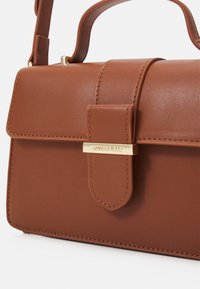 Petit sac à main rectangulaire en cuir marron avec poignée dessus, rabat avant et fermoir doré gravé du logo "ANNA FIELD".