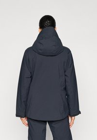 Jachetă impermeabilă de culoare bleumarin închis, cu guler înalt și glugă reglabilă. Dispune de mâneci lungi și accente cu Velcro la manșete. Textură netedă.