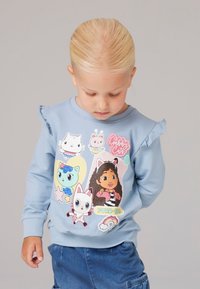 Gaiši zilas krāsas džemperis ar volāniem plecos, rotāts ar krāsainiem multfilmu tēlu apdrukām, tostarp meitenes un dažādu dzīvnieku attēliem.