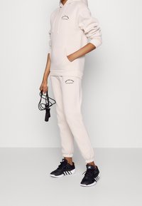 Beige hooded sweatshirt med en framficka och logotyp, matchad med byxor i samma färg som har en logotyp. Svarta träningsskor och hopprep.