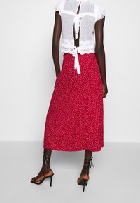 Jupe midi rouge à pois blancs, associée à un haut transparent blanc avec des manches froncées et un lien dans le dos. Talons à lanières noirs visibles.