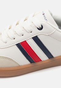 Valge nahast tennis, millel on pruun kummist tall, punaste ja tumesiniste triipudega. Sidumisdisain tekstuurse varbakattega. Logo külje peal.