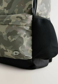 Grau camouflierter Rucksack aus strukturiertem Material, mit einer schwarzen Netzseitentasche und einem runden Logo am Boden. Reißverschluss oben.