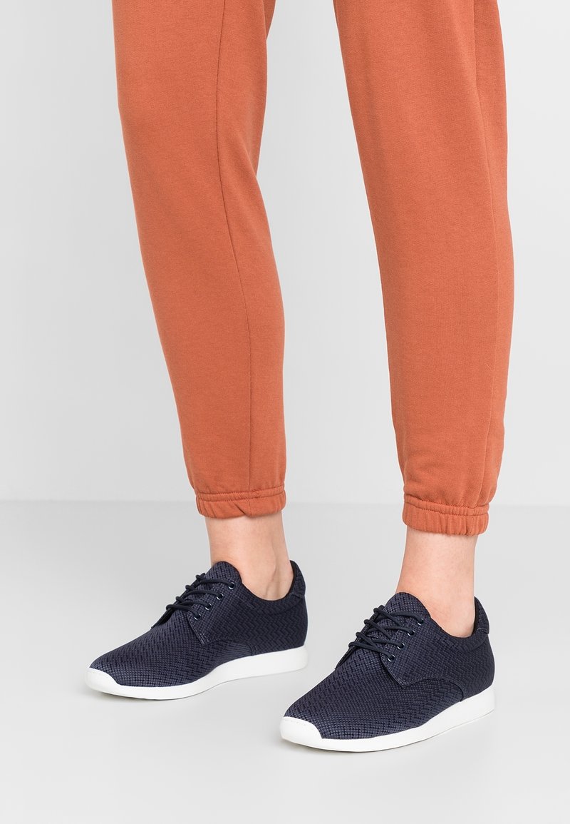 Svarta texturerade sneakers med ett fiskbensmönster, vit sula och matchande snören, tillsammans med orangea tajta sweatpants.