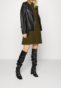 Svart läderbikerjacka över en olivgrön klänning, parat med knähöga svarta slouchy klackstövlar. Släta texturer och solida färger.