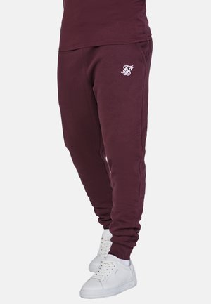 Burgunderfarbene Jogginghosen aus weichem Stoff mit einem weißen Logo auf der Vorderseite, gerippten Bündchen und einem lockeren Schnitt. Kombiniert mit weißen Sneakern.