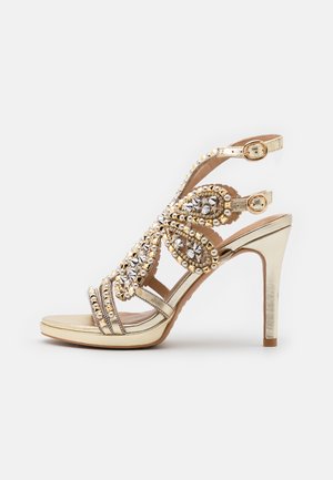 Sandalias de tacón alto doradas con rhinestones decorativos, presentan múltiples correas, un diseño tipo slingback y un acabado metálico suave.