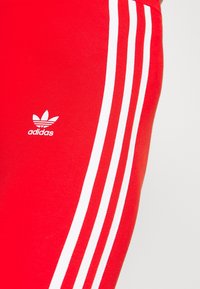 Červené sportovní legíny s třemi bílými pruhy po stranách a malým bílým logem Adidas. Materiál má hladkou texturu.