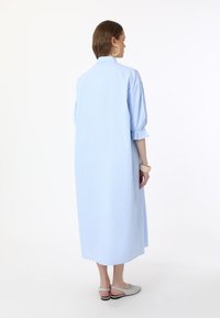 Robe chemise rayée bleu clair avec un col montant, des manches bouffantes et des fentes latérales, fabriquée en tissu léger, allant du genou à la cheville.