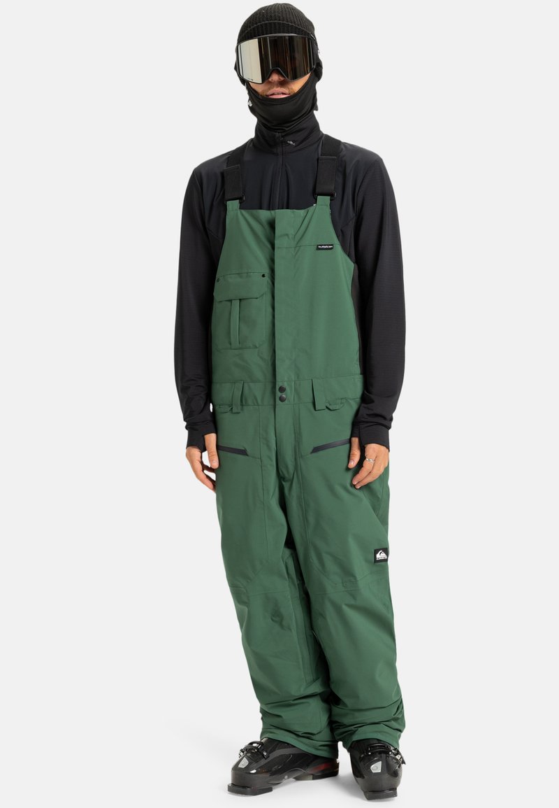 Persoon in groene sneeuwbroek met bretels, zwarte jas, zwarte beanie, skigoggles en skischoenen tegen een effen achtergrond.