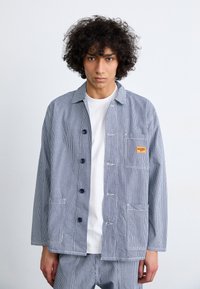 Striped blue and white jacket made of cotton, featuring a button front, two pockets, a small logo patch, and a collar design.