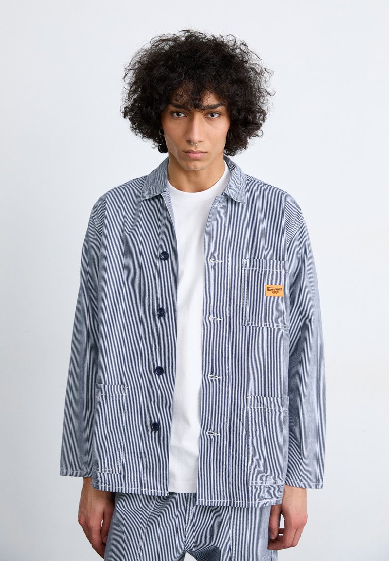Striped blue and white jacket made of cotton, featuring a button front, two pockets, a small logo patch, and a collar design.