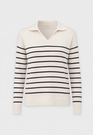 Vrouw met lang blond haar en bril draagt een gestreepte trui en losse beige broek, houdt een grote beige handtas met schouderriem vast.