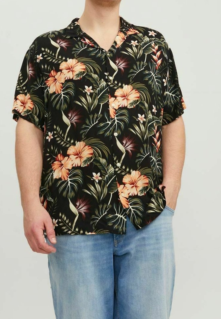 Personne portant une chemise hawaïenne noire à manches courtes avec des fleurs d'hibiscus orange et un jean bleu, debout avec une main dans une poche.