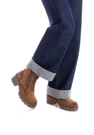 Botas de ante marrón hasta el tobillo con un tacón grueso, detalle de cordones en la parte delantera y cremallera lateral. Combinadas con unos jeans de mezclilla oscura con dobladillo enrollado.