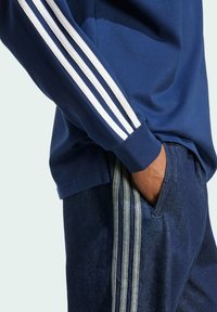 Navy long-sleeve shirt with white three-stripe accents on the sleeves, paired with dark denim pants featuring similar stripe detail.