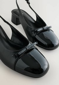 Svarta slingback-skor med låg klack. Glansigt patenterat läder på tåpartiet med en rosettdetalj. Mjuk läderöverdel och foder.
