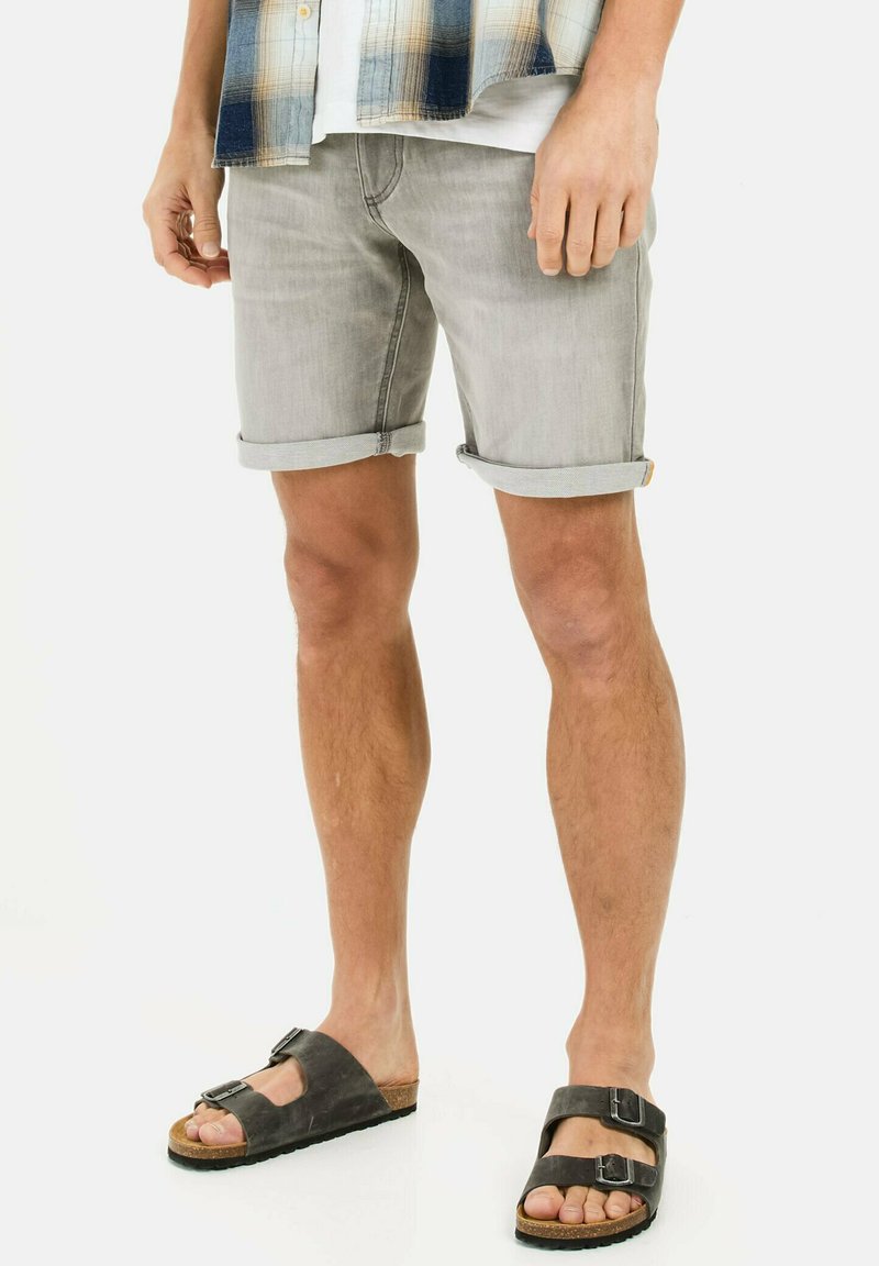 Graue Denim-Shorts mit umgeschlagenen Säumen, kombiniert mit dunklen Sandalen mit doppelten Schnallenakzenten. Das Modell steht mit entspannten Armen an den Seiten.