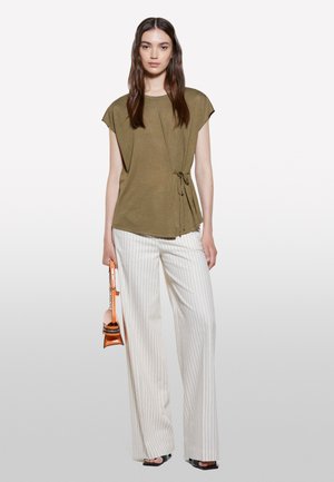 Olive green short-sleeve blouse with a tied waist detail, paired with white vertically striped wide-leg trousers and a small orange handbag.