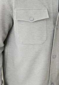 Camisa gris de botones, hecha de un tejido texturizado, con un bolsillo en el pecho con un botón a presión y botones grises a juego en el frente.