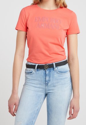 Vrouw die een koraalkleurig Emporio Armani T-shirt draagt, ingestopt in lichtblauwe spijkerbroek met een zwarte riem met een ronde zilveren gesp.
