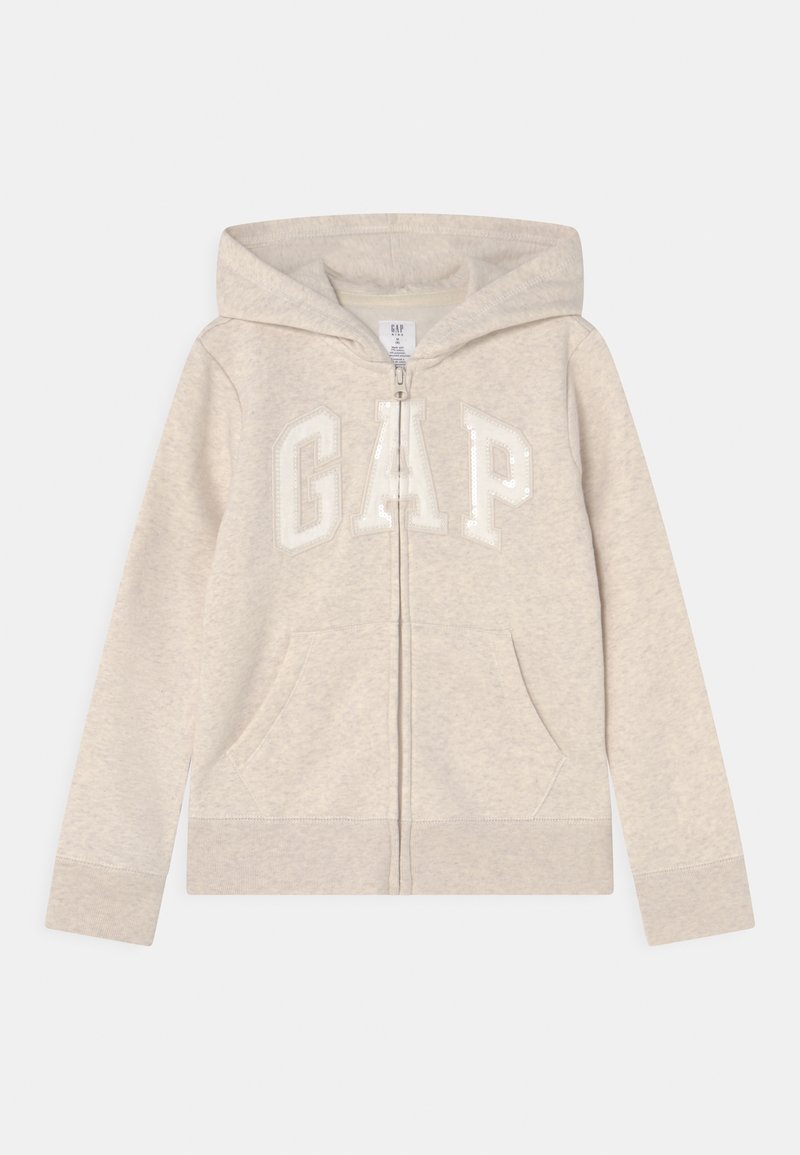 Sweat à capuche beige zippé en tissu doux et texturé. Il possède une capuche et des poches avant, avec l'inscription "GAP" en grosses lettres pailletées blanches sur la poitrine.