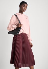Sudadera rosa claro con bolsillo canguro, combinada con una falda burdeos plisada. La modelo lleva un bolso negro de hombro. Texturas suaves.