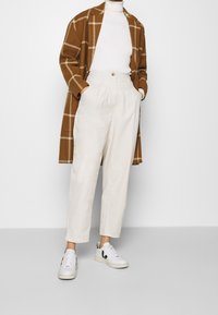 Cappotto a quadri marrone sopra un dolcevita bianco a coste, abbinato a pantaloni plissettati crema e sneakers bianche con dettagli neri.