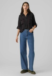 Camicia nera con bottone, dotata di due tasche frontali, abbinata a jeans a gamba larga di un blu chiaro e scarpe chunky verde scuro. Tessuto dalla texture morbida.
