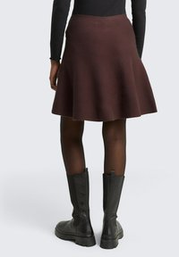 Bruine uitlopende rok van een gestructureerde stof, tot de knie, gedragen over zwarte maillot, gecombineerd met zwarte enkelboots met dikke zool.