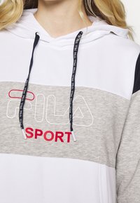 Detailní záběr na bílo-šedou mikinu s černými šňůrkami, se znakem "FILA SPORT" v bílé a červené barvě na hrudi.