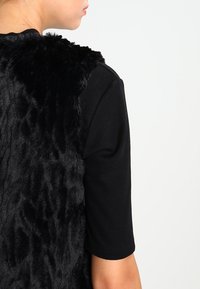 Svart fuskpälsväst med en slät textur, parat med en figurnära svart ribbad kortärmad topp, sedd bakifrån.