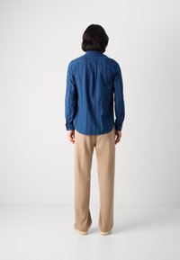 Cămașă din denim cu mâneci lungi, de culoare bleu închis, cu guler butonat și o tijă curbată, purtată cu pantaloni largi beige și pantofi deschiși.