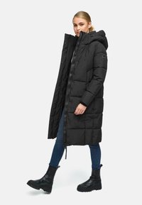 Lang sort puffet frakke med hætte, der har lynlås lukning, elastiske manchetter og store quiltede sektioner. Båret over jeans og sorte støvler.